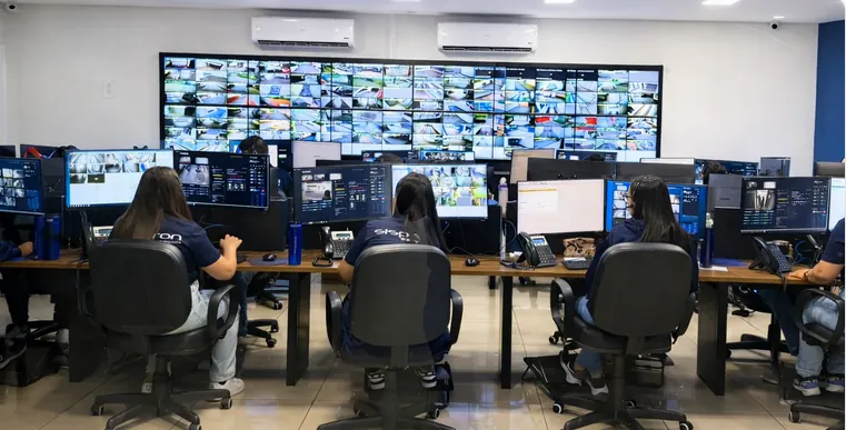 Operadoras na CMS, a Central de Monitoramento Sispron em Santo André, com videowall de câmeras ao fundo